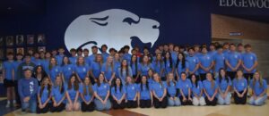 A large group of high school students and one adult, all wearing matching blue shirts, pose for a group photo indoors in front of a wall with a large white lion logo and the word “Edgewood.”.