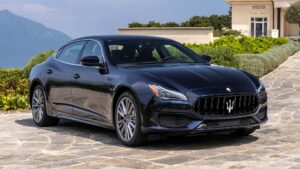 A sleek black Maserati Quattroporte luxury sedan is parked on a stone driveway, its refined presence hinting at premium automotive experience, with manicured greenery and an elegant building—perhaps a campus—in the background on a clear day.