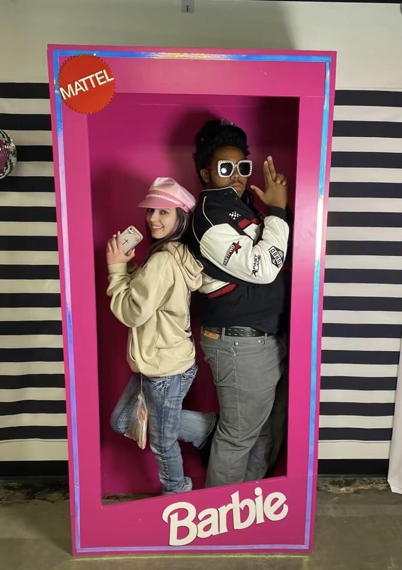 Two people pose back-to-back inside a life-sized Barbie doll box prop. The person on the left wears a pink hat and holds a toy phone, while the person on the right wears sunglasses and makes a peace sign. The background has black and white stripes.