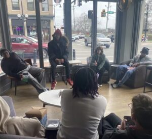 A group of six people sit in a circle at a coffee shop by large windows overlooking a busy street. Some are talking, one person makes a peace sign, and others are listening or on their phones.