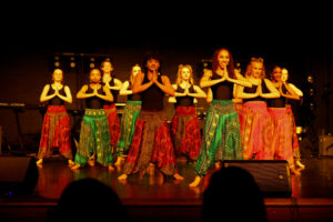 A group of dancers from the School of the Arts perform under stage lights, wearing colorful patterned pants and black tops. Barefoot and in formation, they press their hands together, embodying the spirit of performing arts in Rhythms of the World.