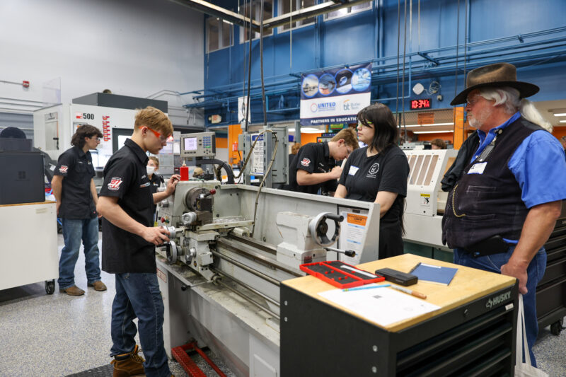 Several people in a workshop observe a young man operating a metal lathe during a machining olympics event. Others watch or work nearby in this educational facility, where students connect and learn hands-on skills with various machines and tools.