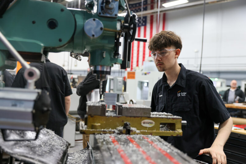 A young man wearing safety glasses operates a milling machine in a workshop, surrounded by metal shavings during the Machining Olympics. Other people and American flags are visible in the background.