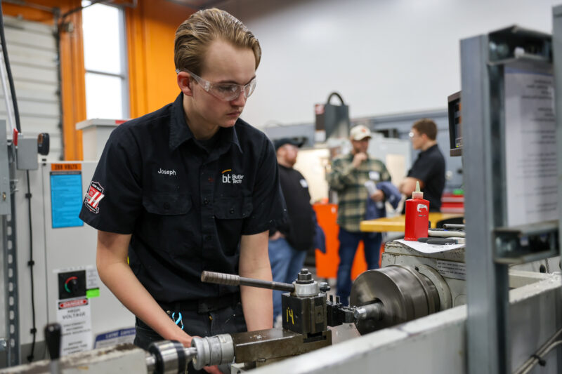 A person wearing safety glasses and a black work shirt operates a lathe machine in a workshop during the machining olympics, with several blurred people in the busy, industrial background.