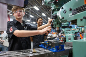 Two students connect during a machining olympics workshop, operating a milling machine together. One adjusts the controls as the other observes and smiles. Both wear black shirts and safety glasses; metal shavings and equipment are visible on the worktable.