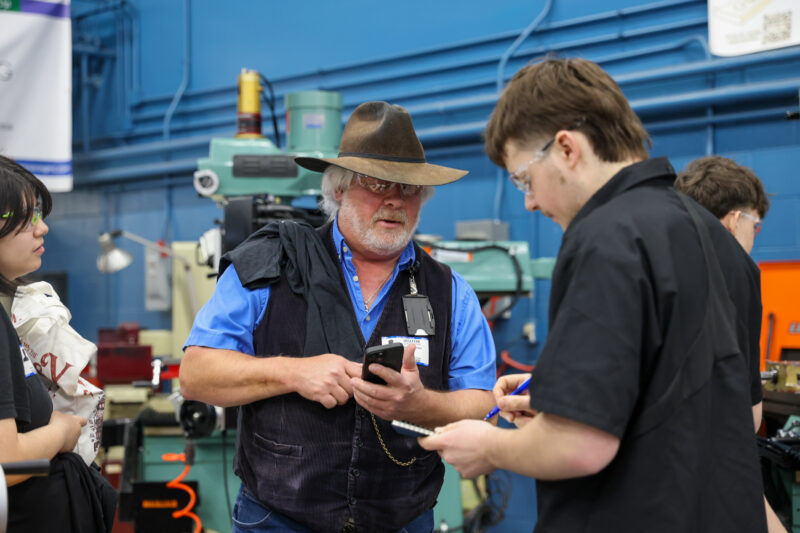 A man in a cowboy hat and vest talks to another man with glasses and a notepad in a workshop during the Machining Olympics, with machinery and tools visible in the background. Other people are also present.