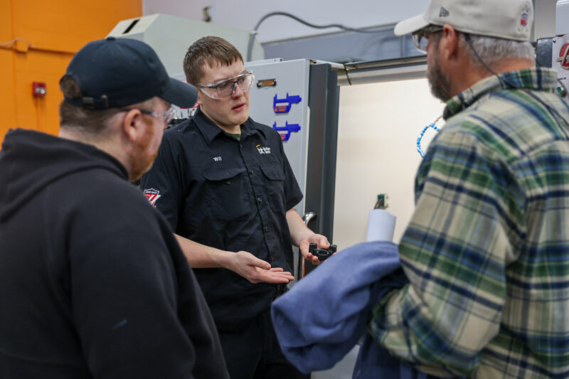 A man in safety glasses and a black work shirt explains something to two men—one in a plaid shirt and cap, the other in a black hoodie and cap—during a machining olympics event in an industrial setting with machinery in the background.