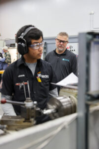 A young man wearing safety glasses and ear protection operates machinery while an older man in glasses observes him, as students connect during a machining olympics event in a workshop or classroom setting.