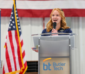 A young woman speaks into a microphone at a podium labeled "Butler Tech," with American and Ohio flags beside her and a large American flag in the background during an Aviation Center ribbon cutting event.