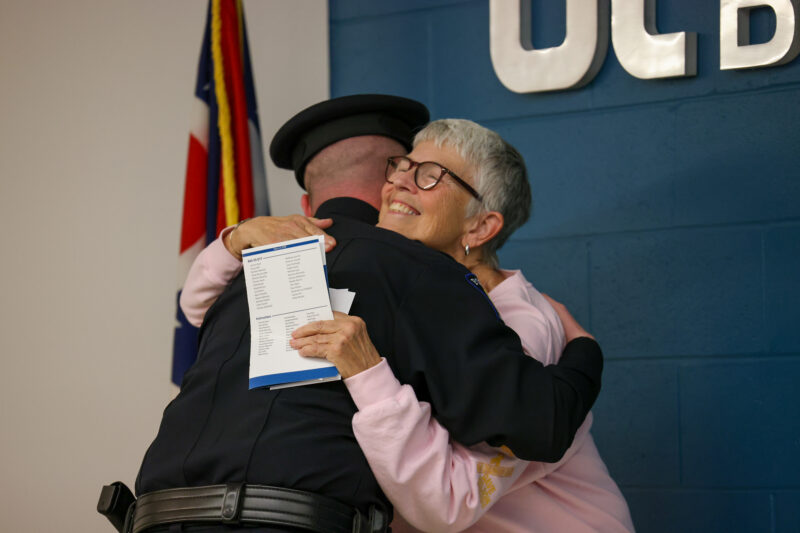 A police officer in uniform, honored with the Legacy of Service, hugs an older woman with short gray hair and glasses, who is smiling and holding papers. They stand indoors near a blue wall with metallic letters and a flag visible.