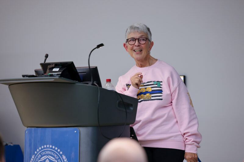 An older woman with short gray hair and glasses stands smiling at a podium. Wearing a light pink sweatshirt, she holds a microphone—ready to present the Sgt Brian Dulle Memorial Award, honoring a Legacy of Service at Butler Tech Police Academy.