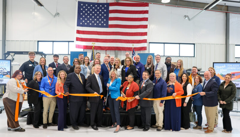 A large group of people pose and smile for a photo at an Aviation Center ribbon-cutting ceremony in front of a large American flag inside an industrial building, celebrating workforce achievements with a long orange ribbon and scissors.
