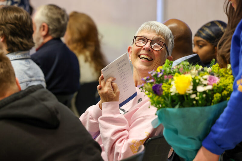 An older woman with short gray hair and glasses smiles joyfully while holding a program at the Butler Tech Police Academy event. She sits among others as a vibrant bouquet is handed to her, honoring the Legacy of Service with the Sgt Brian Dulle Memorial Award.