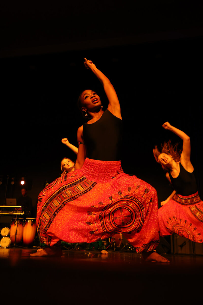 Three dancers from the School of the Arts in colorful, patterned red pants and black tops perform energetically on stage, one with an arm raised and expressive face, under warm lighting at Rhythms of the World with musical instruments in the background.