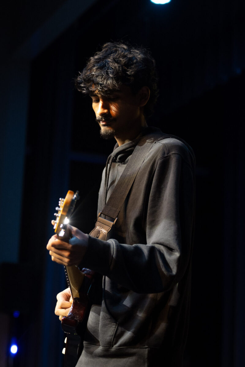 A man in a dark hoodie plays an electric guitar under stage lighting, focused on his performance with a dark background behind him, capturing the spirit of the performing arts at School of the Arts.