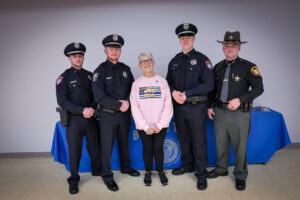 Five people pose for a photo: four police officers in uniform and a smiling older woman in a pink sweatshirt, standing indoors by a blue table with the police emblem, celebrating the Legacy of Service honored by the Sgt Brian Dulle Memorial Award.