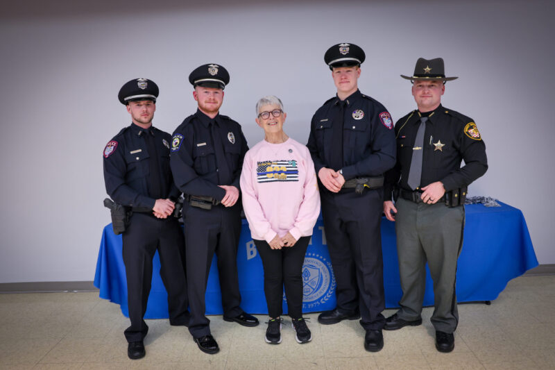 Five people pose for a photo: four police officers in uniform and a smiling older woman in a pink sweatshirt, standing indoors by a blue table with the police emblem, celebrating the Legacy of Service honored by the Sgt Brian Dulle Memorial Award.