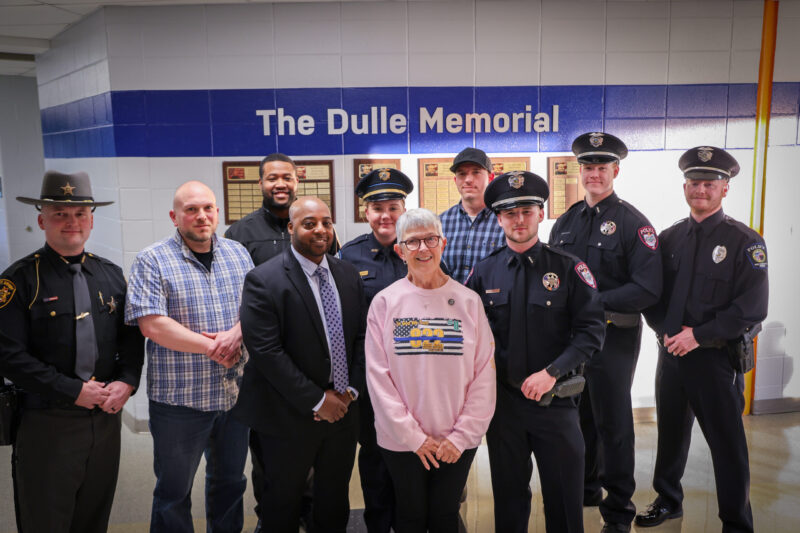A group of nine men and one woman, including police officers in uniform and others in civilian clothing, stand together smiling in front of a wall labeled 