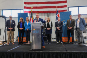 A group of nine people in business attire stand on stage with microphones and banners reading "Butler Tech" behind them, celebrating a Ribbon Cutting. An American flag and Ohio state flag are displayed in the background.
