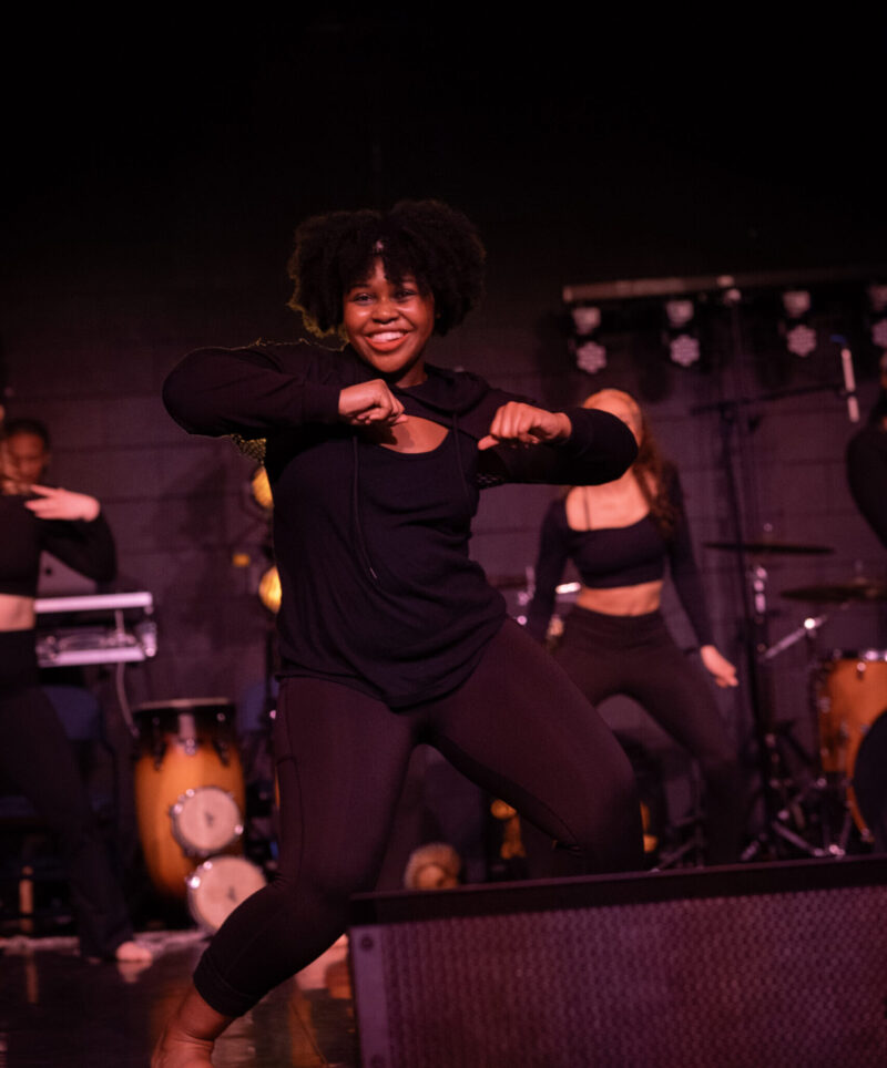 A woman in black clothing performs energetically on stage, smiling, with musicians and percussion instruments in the background. The dimly lit scene glows with purple and yellow lights during the vibrant Rhythms of the World at School of the Arts.