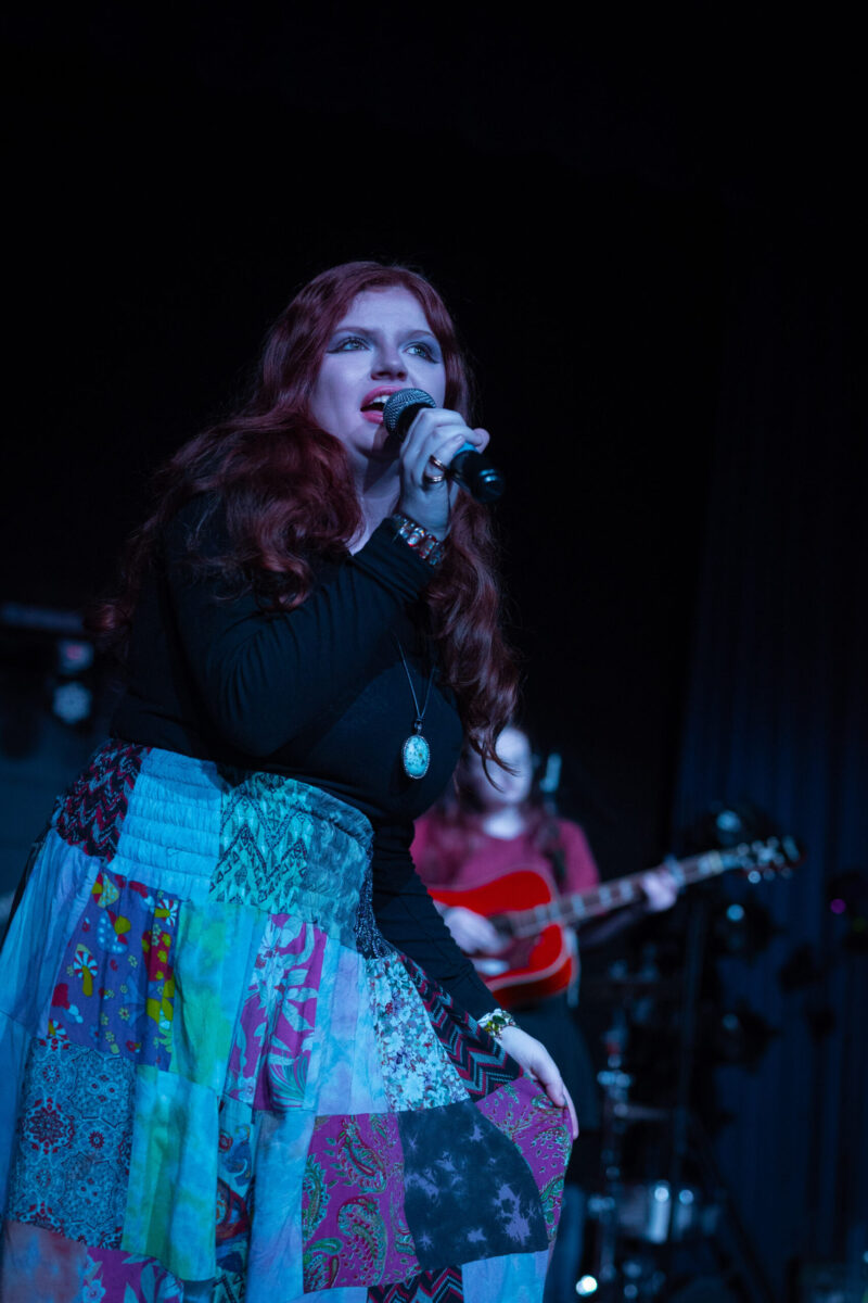 A woman with long red hair, wearing a colorful patchwork skirt and black top, performs on stage at Rhythms of the World. A guitarist from the School of the Arts is seated in the background, playing under blue stage lighting.