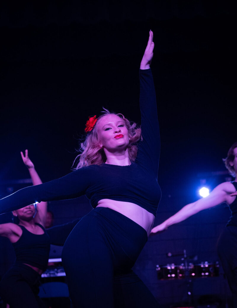 A dancer in a black outfit with a red flower in her hair strikes a dramatic pose on stage during Rhythms of the World at School of the Arts, one arm raised and the other bent under blue lights. Other performers appear in the background.