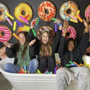 Three people sit in a white bathtub filled with colorful foam sticks, happily throwing them in the air. The background features a wall decorated with large, playful donut graphics.