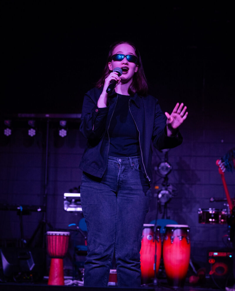 A woman in sunglasses, a black jacket, and jeans performs into a microphone on a dimly lit stage with drums and percussion instruments in the background at the School of the Arts' Rhythms of the World event.