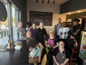 A group of nine smiling people, some standing and some sitting, pose inside a cozy local café with a chalkboard menu behind them decorated for Valentine's Day, celebrating their recent sandwich competition.