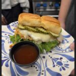 A hand holds a plate with a sandwich made of lettuce, melted cheese, pickles, and a soft bun—just like one from a local café—served with brown dipping sauce. The plate features a blue floral design.