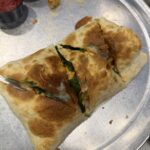A golden-brown calzone filled with vegetables and cheese, sliced into four pieces, sits on a metal tray at a local café. Two small containers of dipping sauces, one red and one yellow, are placed near the top edge.