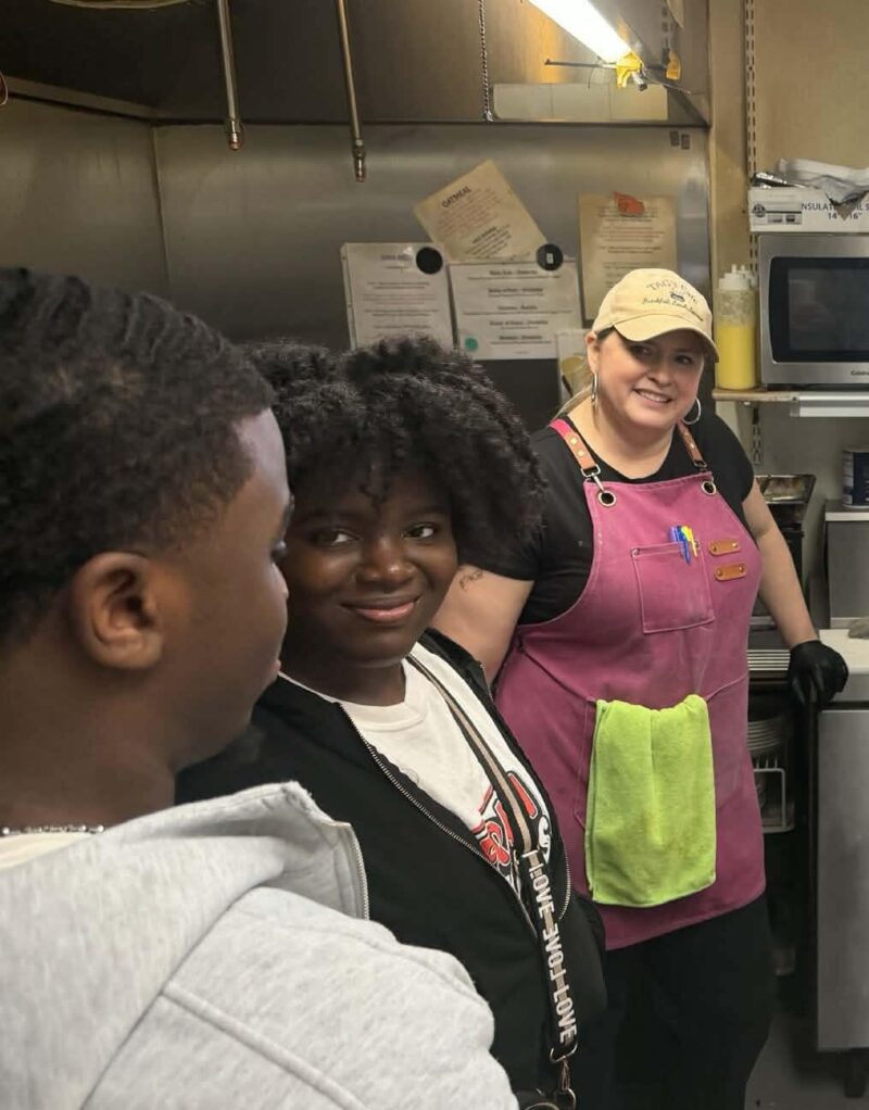 Five people are gathered in a kitchen, smiling and talking. One person, dressed as a culinary student with an apron and hat, chats with friends. Food containers and a used plate hint at their recent sandwich competition.