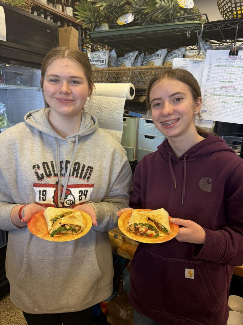Two smiling teenage girls, culinary students, stand indoors holding plates with vegetable-filled wrap sandwiches. Shelves with food and supplies are visible in the background.