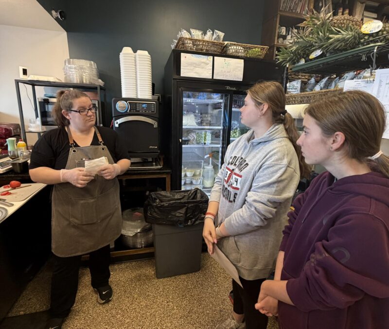A woman in an apron and gloves speaks to two Culinary students in casual clothing inside a kitchen area, with shelves, a fridge, and food containers visible in the background as they prepare for a sandwich competition.