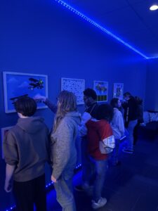 A group of people stand in a dimly lit blue room, looking at and interacting with framed displays on the wall. Blue LED lights line the walls and ceiling, creating a vibrant atmosphere.