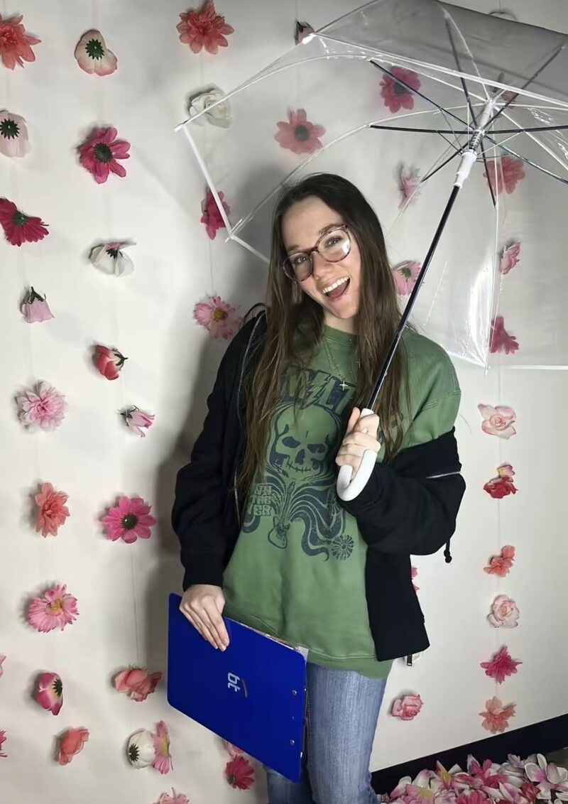 A smiling person with glasses holds a clear umbrella and a blue folder, standing in front of a white wall decorated with pink and red artificial flowers. They wear a green graphic t-shirt, black jacket, and jeans.