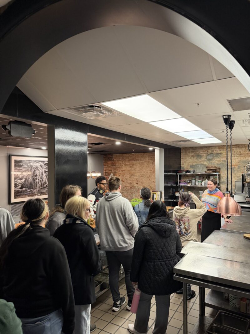 A group of people stand in a restaurant kitchen, listening to a woman holding a paper and speaking. The kitchen has metal counters, brick walls, and overhead lighting.