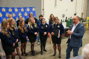 A man speaks to a group of young women and men in blue jackets and black skirts or pants, likely Talawanda–Butler Tech FFA students, standing in an indoor event space with a blue banner highlighting leadership in the background.