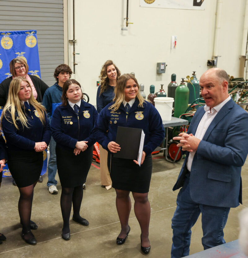 A man speaks to a group of young women and men in matching blue jackets and black skirts or pants, standing in an industrial room with equipment and a Talawanda–Butler Tech FFA banner, highlighting leadership and teamwork.