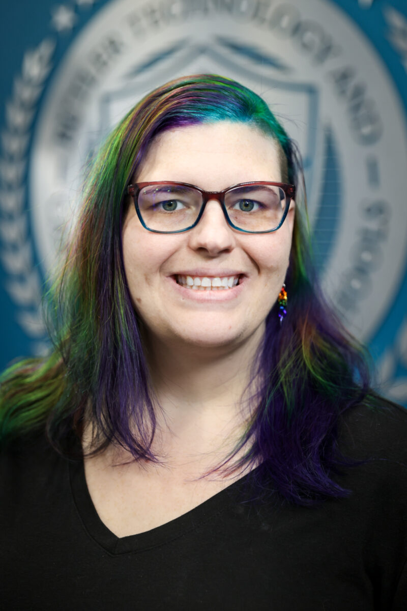 A smiling person with glasses and long hair dyed in green, blue, and purple stands in front of a blurred blue background featuring a circular Board of Education emblem.