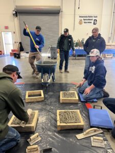 A group of people, demonstrating leadership, work indoors on a concrete project. Some pour concrete into wooden molds while others smooth the surface with trowels. Construction tools and materials are arranged on the floor.