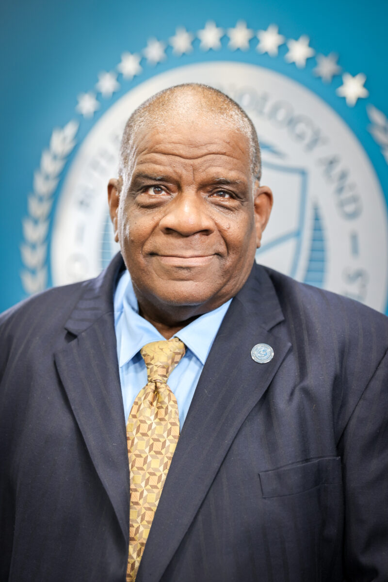 A man in a dark suit, patterned yellow tie, and light blue shirt stands in front of a blurred Board of Education seal, smiling slightly at the camera.