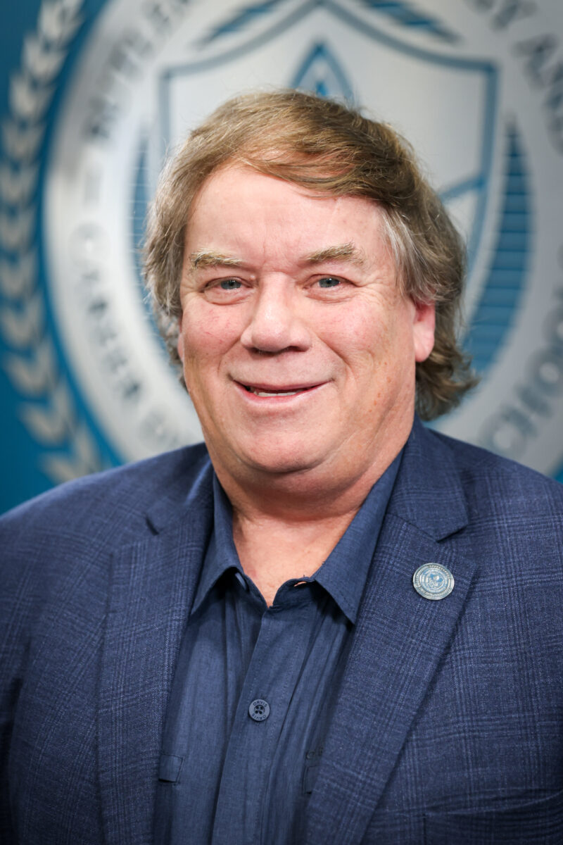 A middle-aged man with light brown hair, wearing a blue blazer and a navy shirt, smiles at the camera. He has a Board of Education pin on his lapel and stands in front of a blue and white emblem background.