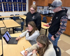 Four students in a classroom collaborate on a project for Talawanda–Butler Tech FFA; two are seated at a table with a laptop and notebook, another stands watching, and one holds a phone. Award banners hang on the wall, reflecting leadership and achievement.
