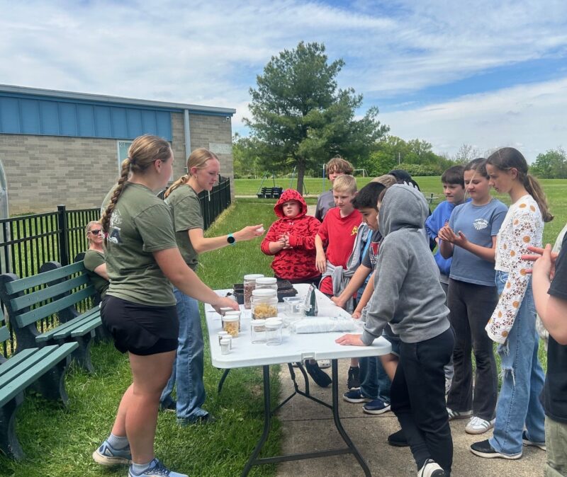 A group of kids and two adults gather around a table outdoors, engaging in an interactive activity with various items. Demonstrating leadership, some children closely observe while others stand nearby on a sunny day.