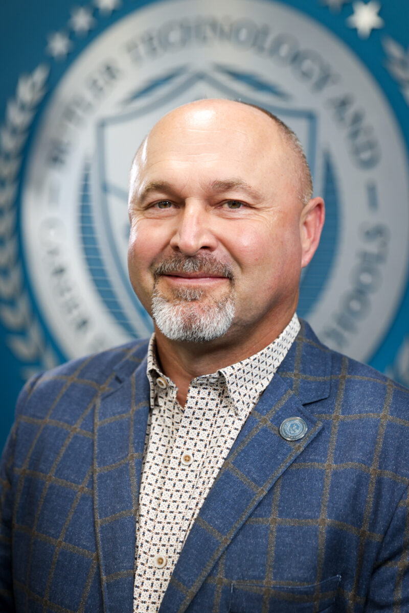 A middle-aged man with a bald head, goatee, and mustache is wearing a blue checkered blazer and patterned shirt, standing before a blue wall with a blurred Board of Education emblem in the background.
