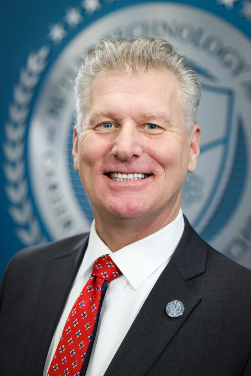 A smiling man with short, gray hair is wearing a dark suit, white shirt, and red patterned tie, standing in front of a blue background with a blurred Board of Education seal.