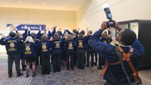 A group of people in matching blue jackets with state and chapter names, including Talawanda–Butler Tech FFA, stand with their backs to the camera, raising one arm in a display of leadership as a photographer captures the moment near an "EXPO HALL" sign.