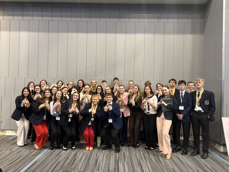 A large group of smiling young people, dressed in business attire with name tags and medals, pose together indoors against a gray paneled wall, some making celebratory hand signs after earning DECA State Honors.