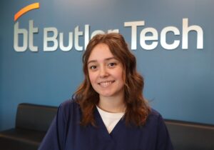 A young woman in navy blue scrubs smiles at the camera, standing in front of a blue wall with the "Butler Tech" logo in large silver letters—a great option for seniors exploring 1-year and 1-semester programs.